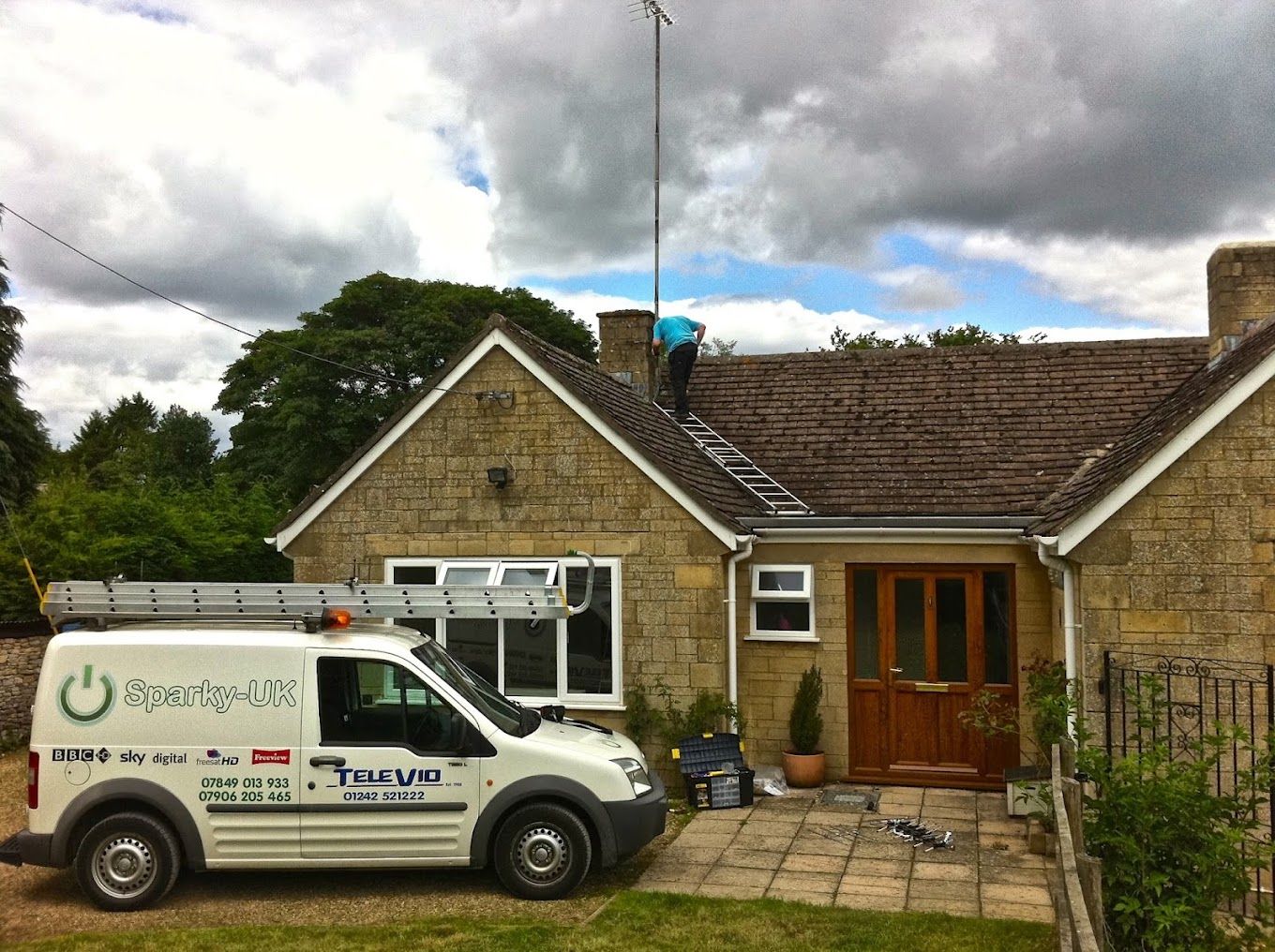 Working on a TV aerial at the chimney of a Cotswold stone bungalow, with the Sparky-UK van parked outside